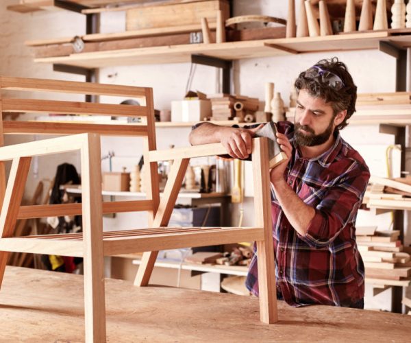 Serious furniture designer carefully sanding a chair frame that he is busy manufacturing in his woodwork studio, with shelves of wooden items behind him
